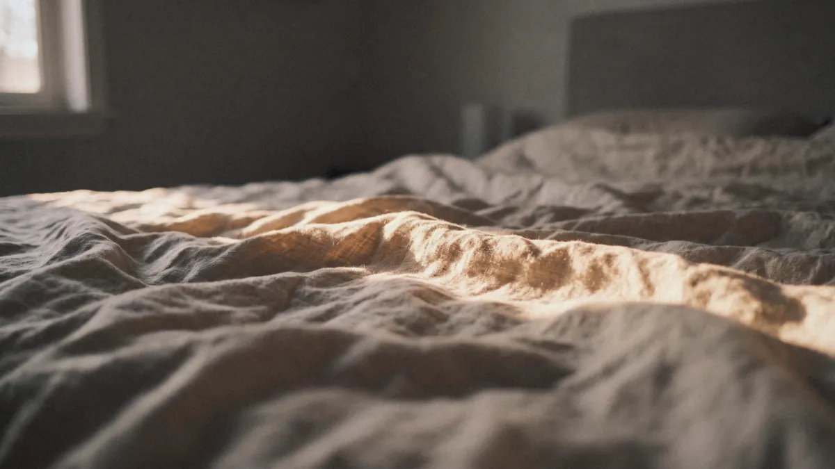 Letto con lenzuola di lino stropicciate attraversate da una lama di luce dorata della sera, come un paesaggio di colline e valli